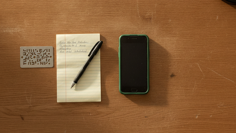 Schreibtisch von oben: ein Smartphone mit grüner Hülle, ein Notizblock mit handschriftlichen Wörtern, ein Kugelschreiber und eine Metallplatte mit eingestanzten Zeichen