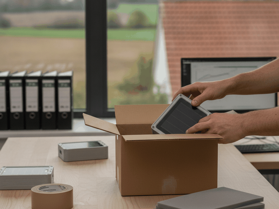 Hände verpacken ein vorkonfiguriertes Sensorgehäuse mit Solarmodul in einen Versandkarton